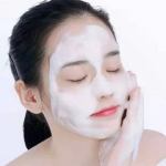 Woman applying facial cleanser foam, eyes closed.