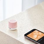 lexon mino+ mini bluetooth speaker pantone rose pink Pink speaker beside smartphone with playlist screen.