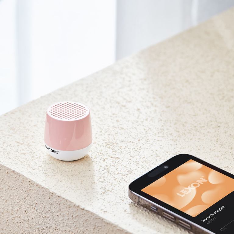lexon mino+ mini bluetooth speaker pantone rose pink Pink speaker beside smartphone with playlist screen.