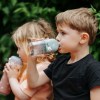Two children drinking from reusable water bottles outdoors