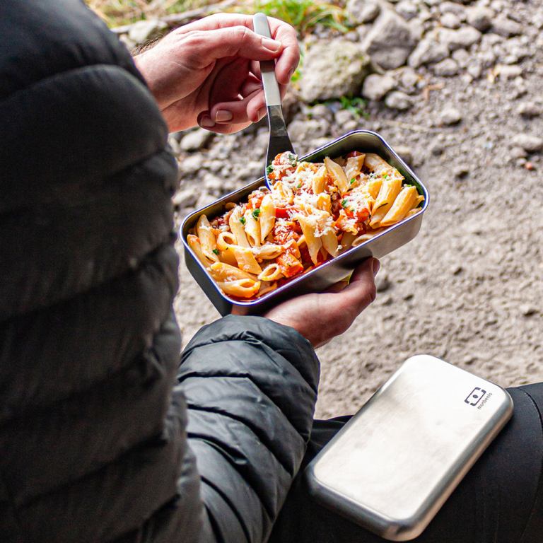 Person eating pasta from stainless steel lunchbox outdoors